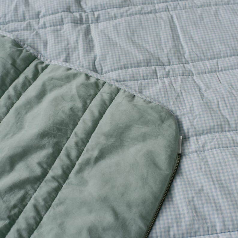 May include: A close-up of a quilted blanket with a light blue and white gingham pattern. The blanket has a solid sage green border with vertical stitching and a silver zipper.