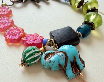Puede incluir: Una pulsera multicolor con cuentas rosas en forma de flor, cuentas verde lima translúcidas, una cuenta cuadrada negra, un dije de elefante turquesa, un pequeño dije de elefante de bronce y una cuenta a rayas verdes y blancas con detalles metálicos.