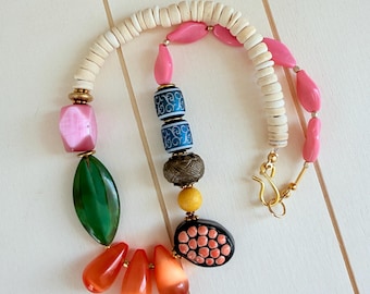 Puede incluir: Un collar que presenta una mezcla de cuentas de disco blancas, cuentas de lágrima rosas, una cuenta rosa facetada grande, una cuenta ovalada verde, tres cuentas de lágrima naranjas, dos cuentas cilíndricas azules con patrones, una cuenta de bronce texturizada, una cuenta amarilla y una cuenta negra con acentos circulares rosas.