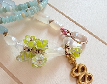 Puede incluir: Un collar de cuentas con abalorios en azul claro, blanco translúcido y transparente, decorado con pequeñas flores de cristal en color verde lima, una piedra transparente ovalada facetada y un colgante de serpiente de metal dorado.