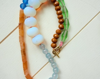 Puede incluir: Un collar de cuentas multicolor que presenta una mezcla de cuentas redondas, facetadas y cilíndricas en tonos azul, blanco, naranja, marrón y verde. El collar se asegura con un cierre de palanca de metal dorado y está ensartado en un cordón rosa.