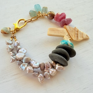 May include: A unique bracelet with a mix of natural materials. It features freshwater pearls, polished stones, and colourful gemstones. A gold-tone clasp and chain secure the bracelet, which also includes a carved bone bird. The bracelet is approximately 20 cm in length.
