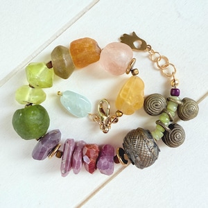 May include: A multi-colored bracelet featuring an assortment of raw, irregular gemstone beads in shades of green, yellow, orange, pink, and purple, interspersed with metallic spiral charms, a small hamsa hand charm, and a gold-toned lobster clasp.