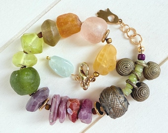 Puede incluir: Una pulsera multicolor que presenta un surtido de cuentas de piedras preciosas en bruto e irregulares en tonos verde, amarillo, naranja, rosa y morado, intercaladas con dijes de espiral metálicos, un dije de mano de hamsa y un cierre de mosquetón dorado.