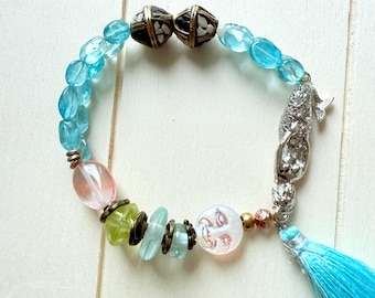Puede incluir: Una pulsera de cuentas con abalorios translúcidos en azul claro y rosa, dos cuentas de color marrón oscuro con patrones y un dije de sirena plateado. La pulsera incluye pequeños espaciadores color bronce, una cuenta blanca y una borla azul claro.