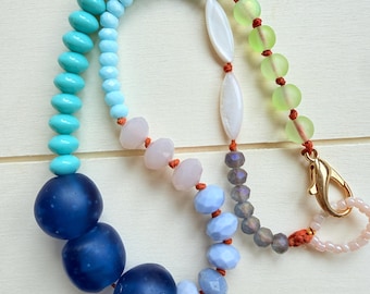 Puede incluir: Un collar que presenta una variedad de cuentas, incluyendo esferas grandes de color azul oscuro mate, cuentas facetadas de color azul claro y turquesa, esferas translúcidas de color verde lima, cuentas blancas ovaladas alargadas y pequeñas cuentas grises facetadas en un cordón naranja.