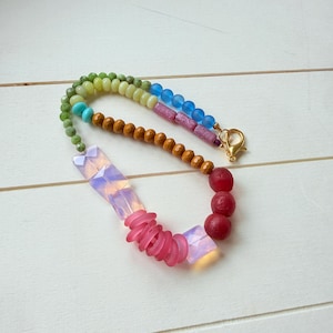 May include: A multi-colored beaded necklace featuring small green, blue, and brown beads, two iridescent rectangular beads, a cluster of pink disc-shaped beads, and three round red beads, finished with a gold-toned lobster claw clasp.