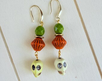 Puede incluir: Un par de pendientes colgantes con ganchos dorados, cada uno con una cuenta verde facetada, una cuenta naranja acanalada y una cuenta en forma de calavera de color amarillo pálido en la parte inferior. Las calaveras tienen cuencas oscuras.