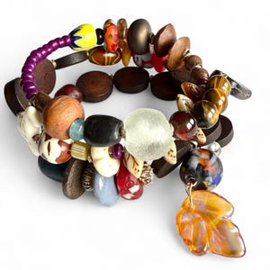 Könnte beinhalten: Mehrstrang-Armband mit einer Vielzahl von Perlen in verschiedenen Formen und Farben. Das Armband umfasst Holzperlen, Glasperlen und dekorative Anhänger, mit einem auffälligen bernsteinfarbenen Blatt-Anhänger.