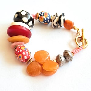 May include: A colorful beaded bracelet featuring a variety of shapes and patterns. The bracelet includes black and white polka dot beads, red and orange beads, and a gold-tone clasp. The beads are strung together with pink and gold accents.