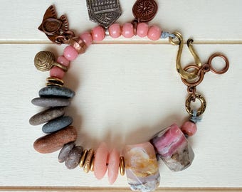 Puede incluir: Una pulsera con cuentas rosas, piedras de río lisas en gris y marrón, y dos grandes colgantes de piedra facetada multicolor. La pieza incluye dijes de cobre y latón, como un escudo y una brújula, con un cierre de gancho de latón.