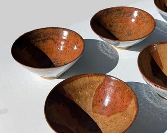 May include: Six small ceramic bowls with a brown and blue glaze. The bowls are arranged in a semi-circle on a white surface.