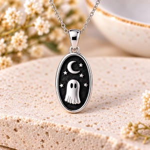 May include: A silver oval pendant necklace featuring a polished ghost figure centred on a dark, textured background. Above the ghost is a crescent moon surrounded by four small stars. The pendant hangs from a delicate silver chain link.