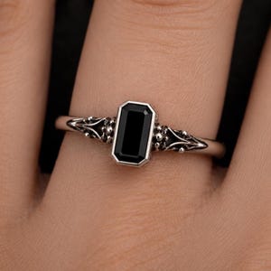 May include: A silver ring featuring a rectangular, emerald-cut black stone set in a bezel. The band displays intricate, textured metalwork with small spherical accents extending from the central setting along the sides of the finger.