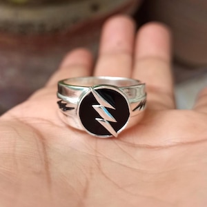 Könnte beinhalten: Ein silberner Ring mit einem schwarzen, kreisförmigen Emblem, das einen weißen Blitz zeigt. Der Ring hat ein dickes Band, wahrscheinlich aus Metall. Dieser Ring ist ein Accessoire zum Thema Superhelden.