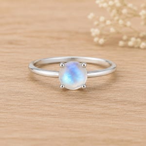 May include: A silver ring featuring a single round, faceted moonstone set in a four-prong setting. The gemstone displays a translucent appearance with a soft blue iridescent glow. The band is smooth, polished, and circular in design.