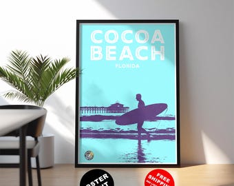 May include: A framed poster print featuring a turquoise background with the words "COCOA BEACH FLORIDA" in white. A purple silhouette of a surfer walking towards the ocean with a surfboard is in the foreground. A pier is in the background.