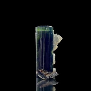 May include: A dark green and blue tourmaline crystal with a clear quartz formation attached. The tourmaline has vertical striations and sits on a dark, rough base. The background is black.