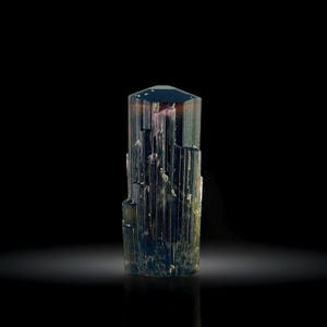 May include: A multi-colored tourmaline crystal with a hexagonal top. The crystal has vertical striations and a gradient of colors, including blue, green, pink, and brown. The gemstone is set against a black background.