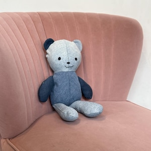 May include: A handmade blue and gray teddy bear with a smiling face, sitting on a pink velvet chair. The bear has dark blue ears, arms, and legs, and a lighter gray body. The chair has a curved back and armrests.