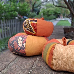 May include: Three fabric pumpkins in various autumn-inspired patterns and textures. One pumpkin features a leaf print, another has a geometric design, and the third displays vertical stripes in shades of orange, yellow, brown, and green.