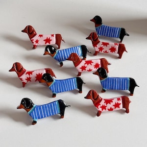 Puede incluir: Una colección de figuras de perro salchicha. Algunos llevan suéteres a rayas azules, mientras que otros tienen cuerpos rosas con patrones de estrellas rojas. Los perros tienen rasgos marrones y negros, y están dispuestos sobre una superficie blanca.