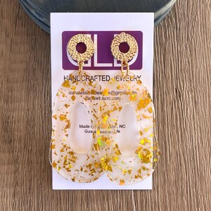 Puede incluir: Un par de pendientes de resina transparente y dorada con un dije circular dorado. Los pendientes tienen forma ovalada y un acabado de purpurina dorada. Los pendientes est&aacute;n hechos a mano y se fabrican en los Estados Unidos.
