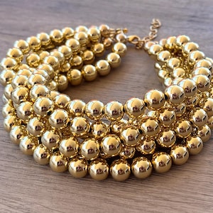 May include: A multi-strand necklace composed of numerous polished gold-tone spherical beads. The necklace features several rows of these reflective, round beads arranged in a dense, layered formation, secured with a gold-tone lobster claw clasp.