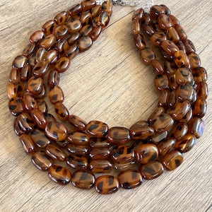 May include: A multi-strand statement necklace featuring polished, oval-shaped beads in a translucent brown and black tortoiseshell pattern. The necklace consists of five layered strands of beads connected by a silver-toned metal clasp and chain extender.