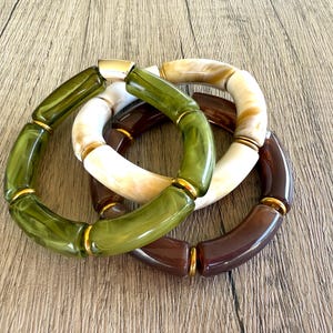 May include: Three stacked bracelets with a mix of colors. One bracelet is olive green, another is white and tan, and the third is a deep brown. Each bracelet has gold-colored metal accents.