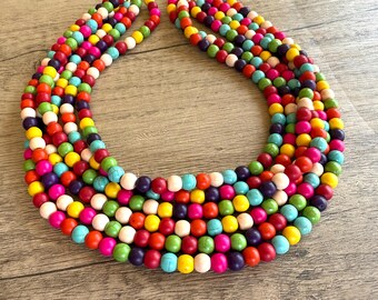 May include: A multi-strand necklace featuring numerous round beads in a vibrant array of colors including turquoise, hot pink, bright yellow, lime green, orange, purple, and white. The beads are arranged in several circular layers of varying lengths.