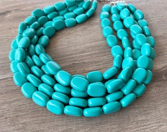 May include: A multi-strand statement necklace featuring five rows of smooth, polished, oval-shaped turquoise beads. The beads are uniform in size and color, creating a vibrant, layered look that connects to a silver-toned metal clasp at the top.