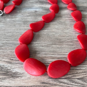 Pode incluir: Um colar vermelho feito de contas grandes de forma oval. As contas são lisas e têm um acabamento ligeiramente brilhante.