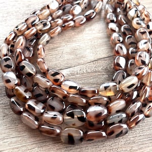 May include: A multi-strand necklace composed of smooth, oval-shaped beads. The beads feature a translucent, light brown base color with dark brown and black mottled, tortoiseshell-like patterns throughout each individual bead surface.