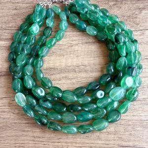 May include: A multi-strand necklace featuring numerous polished, oval-shaped green beads. The beads exhibit varying shades of translucent and opaque emerald green. The strands are gathered at the top with a silver-toned metal clasp mechanism.