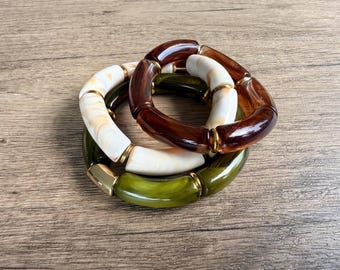 Pode incluir: Um conjunto de três pulseiras elásticas com contas cilíndricas em resina castanho-escura brilhante, creme marmoreado e verde-azeitona. Cada pulseira é acentuada com pequenos espaçadores de metal dourado polido entre as contas lisas.