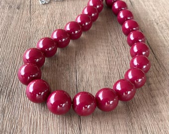 Può includere: Una collana composta da un singolo filo di perline rotonde, lisce e lucide di colore rosso scuro. Le perline sono uniformi per dimensioni e forma, disposte in una linea curva. Una piccola parte di una catenella in metallo argentato è visibile in alto.