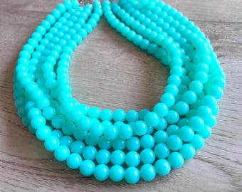 May include: A multi-strand necklace featuring several rows of smooth, round, bright turquoise beads. The beads are uniform in size and color, creating a layered, circular design that connects at the top with a metallic clasp mechanism.