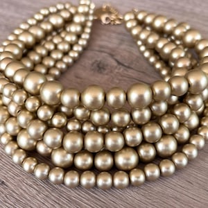 Puede incluir: Collar de cuentas doradas de varias hebras. El collar presenta cuentas redondas de diferentes tamaños, creando un efecto de capas. El cierre es dorado. El collar se muestra sobre una superficie de madera.