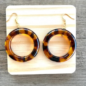 May include: A pair of circular drop earrings featuring a translucent brown and black tortoiseshell pattern. The earrings are suspended from gold-toned metal fishhook wires, creating a classic hoop design with a smooth, polished finish.
