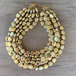 May include: A multi-strand statement necklace featuring numerous polished gold-tone metallic beads in irregular, rounded pebble shapes. The necklace is constructed with several cascading layers of these smooth, reflective beads connected by a gold-tone chain.