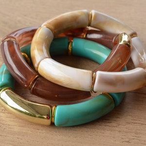 May include: Three stacked bracelets with gold accents. The top bracelet is white with brown accents, the middle bracelet is brown, and the bottom bracelet is turquoise.
