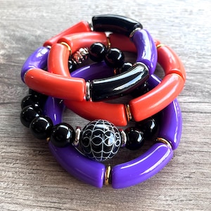 May include: A set of three bracelets featuring a black and white patterned bead, black beads, and colorful plastic tubes in shades of orange and purple.