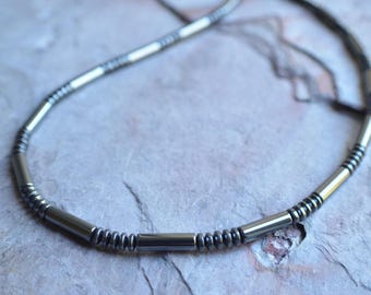 May include: A necklace featuring a repeating pattern of metallic gray cylindrical tube beads and small, round, dark gray disc beads. The beads are strung together in a continuous sequence, creating a sleek, uniform design with a polished finish.