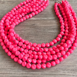 Puede incluir: Un collar rosa brillante de varias hebras con grandes cuentas redondas. El collar tiene un cierre plateado y se presenta sobre una superficie de madera. Las cuentas son de tamaño uniforme y están muy juntas, creando un accesorio llamativo.