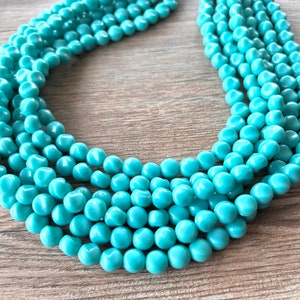 May include: A multi-strand necklace featuring numerous small, smooth, round turquoise-colored beads. The beads are arranged in several parallel rows that curve to form a circular shape, creating a layered appearance with a consistent glossy finish.