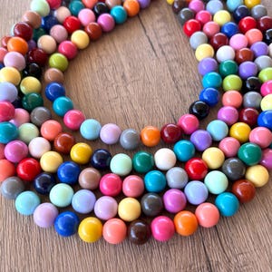 May include: A necklace with multiple strands of colourful, round beads. The beads are in shades of red, pink, orange, yellow, green, blue, brown, and grey. The necklace is displayed on a light brown wooden surface.