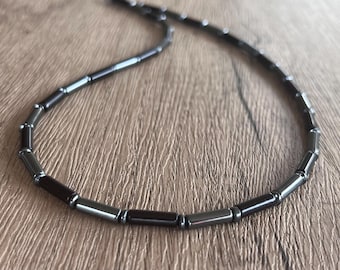 May include: A necklace composed of alternating dark gray and black cylindrical beads. The beads have a smooth, reflective, metallic finish and are strung together in a continuous line, creating a uniform pattern throughout the entire length.