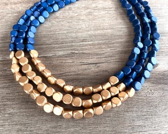 Puede incluir: Un collar de varias hebras con tres filas de pequeñas cuentas cuadradas redondeadas. Las cuentas son de color oro metálico mate y azul real intenso, dispuestas en un patrón de bloques de color donde las cuentas doradas forman la sección central.
