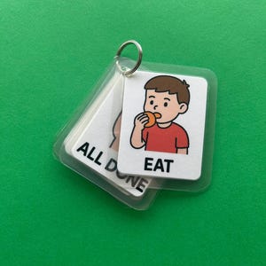 Peut inclure: Un ensemble de cartes de communication visuelle reliées par un anneau métallique. Une carte montre un enfant de dessin animé en train de manger, avec le mot "EAT" en dessous. Une autre carte dit "ALL DONE". Les cartes sont dans des pochettes en plastique transparent.
